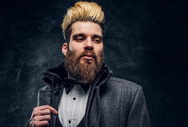 A stylish bearded man dressed in an elegant suit and coat standing against a dark textured wall Dela Cut